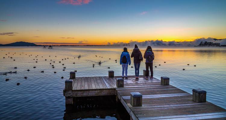 Three people stand on a dock overlooking a misty lake at sunset.