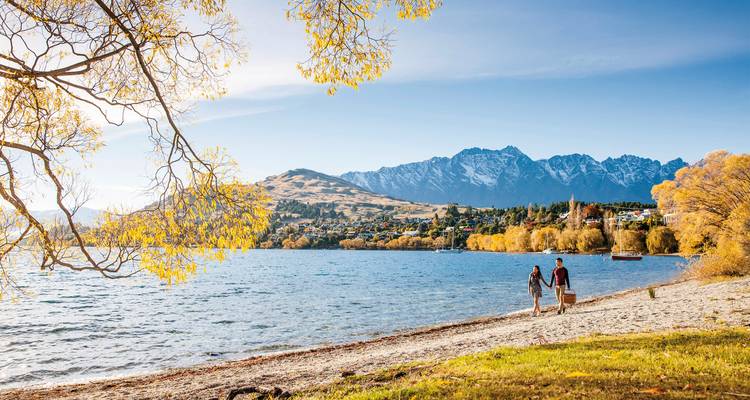 A couple walking along a lakeside beach with mountains.