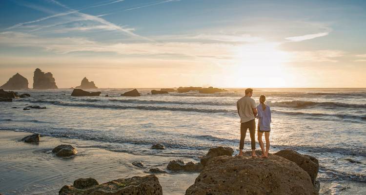A couple standing on rocks overlooking the ocean at sunset.