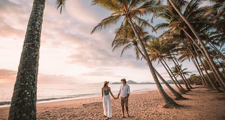 Un couple se tenant la main sur une plage bordée de palmiers au lever du soleil.
