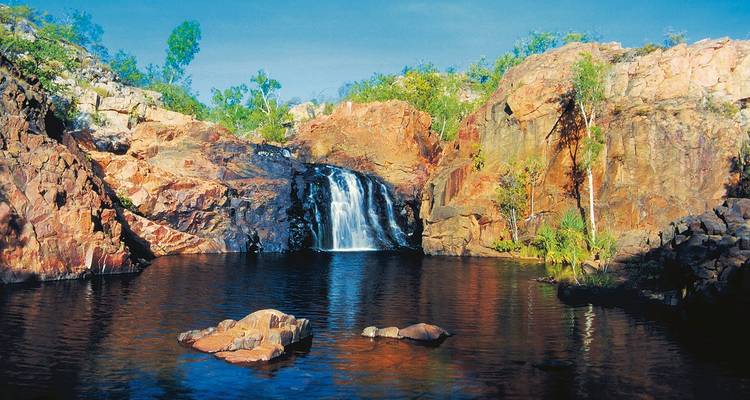 Wasserfall, der über einen felsigen Vorsprung in einen von Steinen und Sträuchern umgebenen Teich fließt.