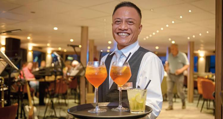 Serveur souriant présentant des boissons sur un plateau dans un cadre intérieur chaleureux.