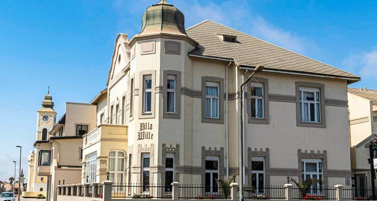 Eine historische Villa im deutschen Kolonialstil mit kunstvoll gestalteten Turmdetails steht unter einem klaren blauen Himmel.