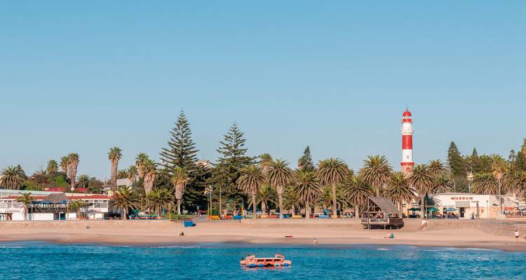 Eine Küstenszene mit palmenumsäumtem Strand und dem rot-weißen Leuchtturm von Swakopmund, der sich dahinter erhebt.