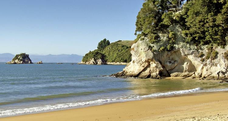 Sonniger Strand mit goldenem Sand und sanften Wellen, türkisfarbenem Wasser, kleinen vorgelagerten Eilanden und bewaldeten Landzungen unter klarem blauen Himmel.