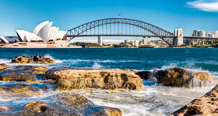 Klassiek ansichtkaartplaatje van het Sydney Opera House en de Harbour Bridge achter kolkende golven op rotsige kust.