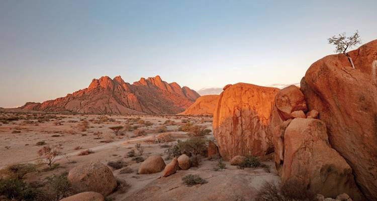 Rotgranit-Gipfel des Spitzkoppe, die in warmem Abendlicht über einer Wüstenebene mit verstreuten Felsblöcken leuchten.