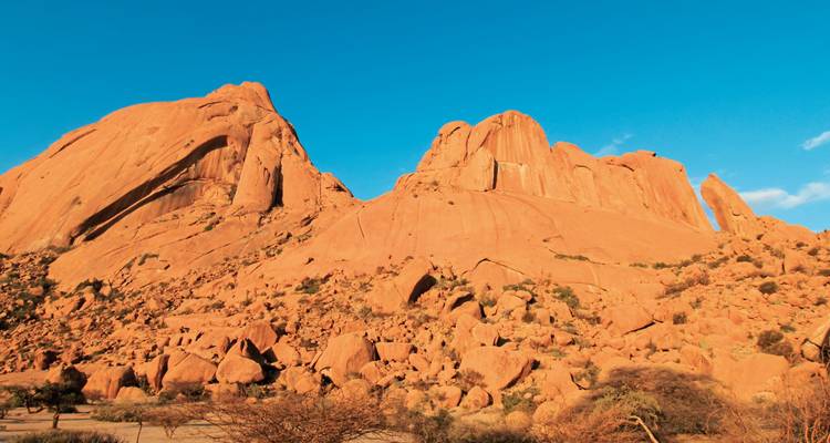 Strahlend blauer Himmel über ragenden orangefarbenen Granitformationen und Feldermeeren in der namibischen Wüste.