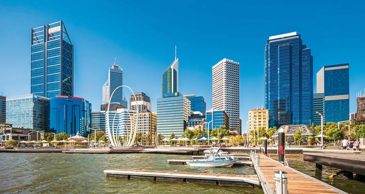 Moderne Skyline aus Glastürmen entlang eines sonnigen Wasserfronthafens mit Booten und Strandpromenade.