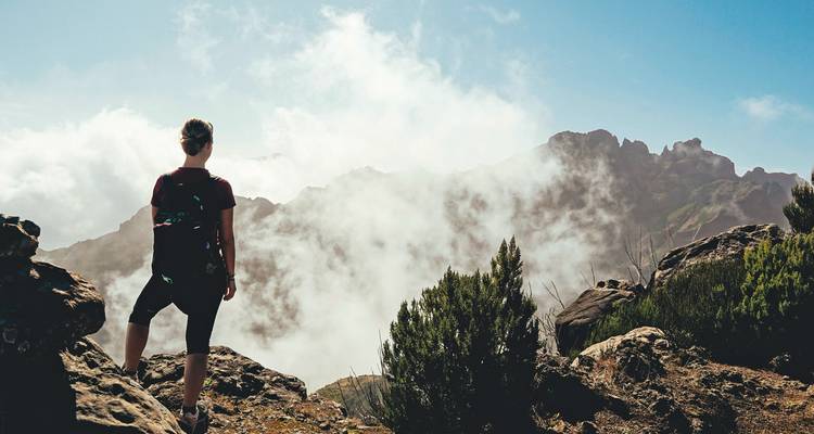 Ein Wanderer steht auf einem felsigen Grat über Wolken mit gezackten Bergspitzen, die teilweise durch Nebel verborgen sind.