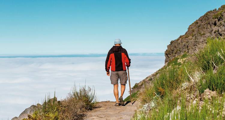 Wanderer mit Wanderstab spaziert entlang eines hochgelegenen Bergpfads über einem Wolkenmeer.