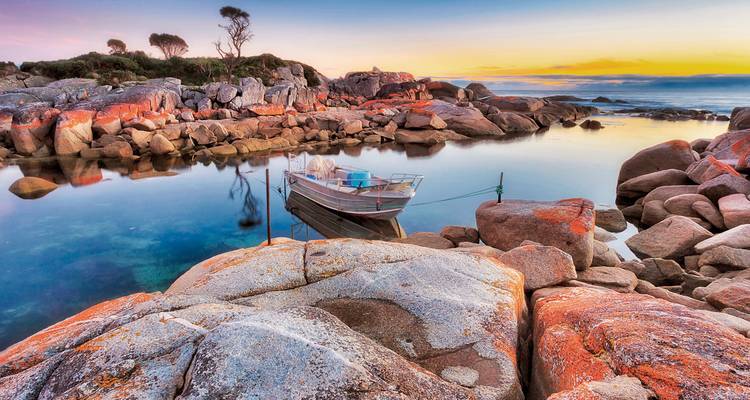 Farbenfrohe Granitfelsen und ein kleines Boot in einer kristallklaren Bucht während des Sonnenaufgangs in Tasmanien.