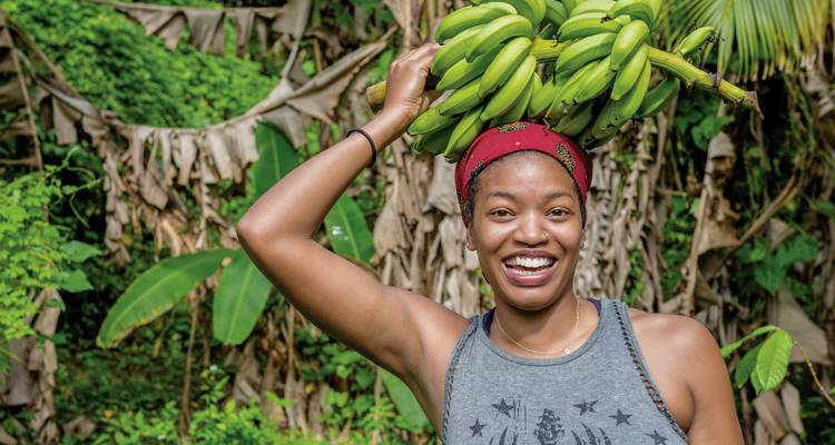 Femme souriante tenant une grappe de bananes vertes dans un cadre tropical.