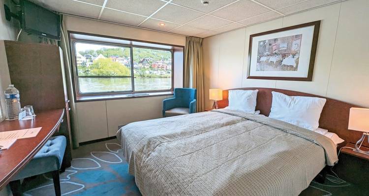 Chambre confortable avec une grande fenêtre sur un bateau de croisière fluviale, vue extérieure sur des bâtiments.