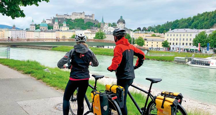 Two cyclists with bikes overlooking a river and cityscape.