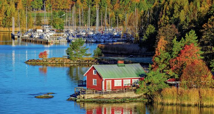 A red cabin by the water with sailboats in a marina surrounded by autumn trees.