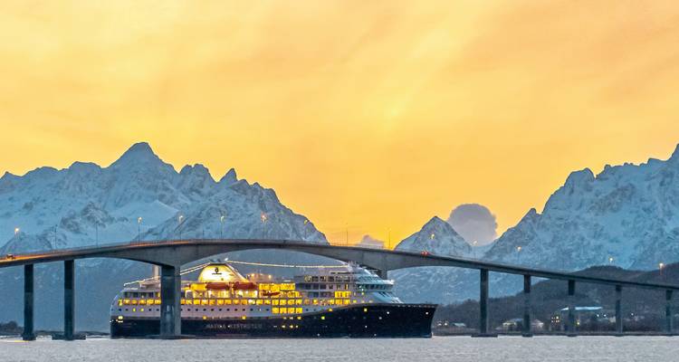 Navire de croisière passant sous un pont avec des montagnes au coucher du soleil.