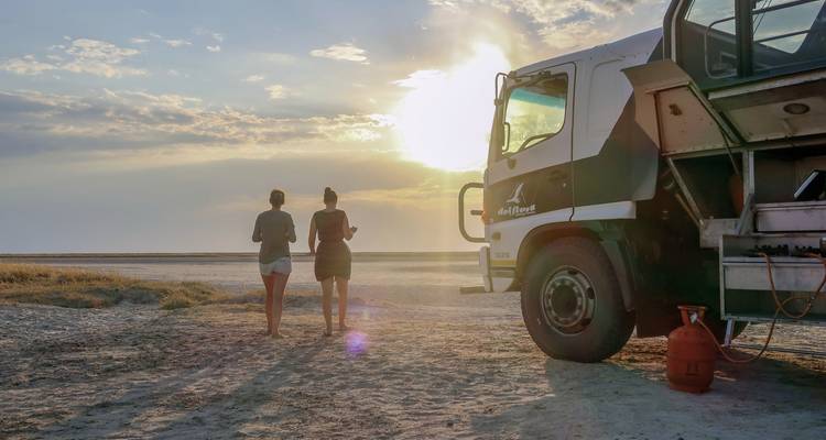 Two people walking near a large truck in a dry landscape, sun setting.