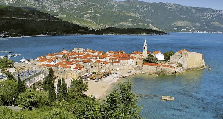 Ville côtière avec des bâtiments aux toits rouges et une petite plage, entourée de montagnes.