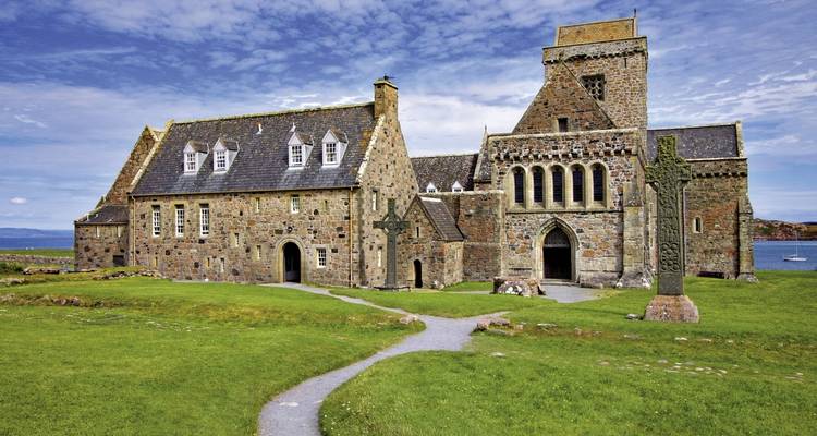 Historic stone building with arched windows and a stone cross in a grassy area.