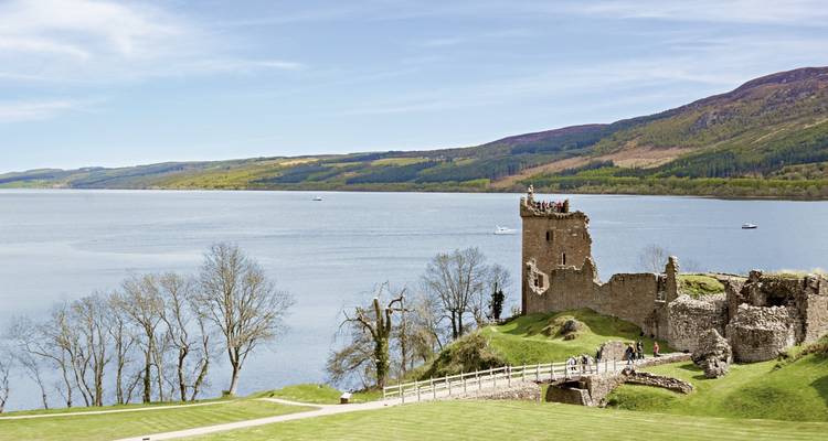 Burgruine an einem See mit Grün und Bergen im Hintergrund.