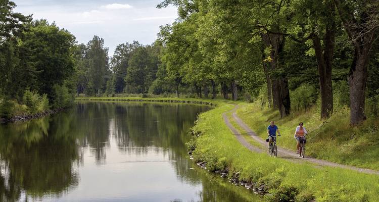 Two people cycling along a canal with lush green surroundings.