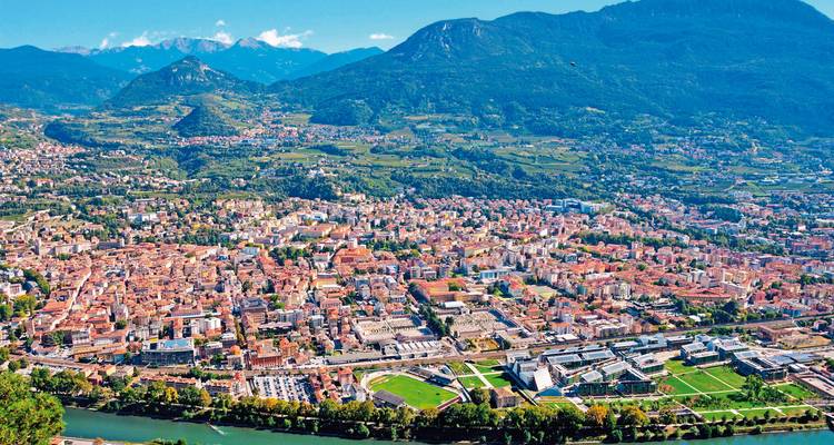 Luftaufnahme einer historischen Stadt mit roten Dächern, eingebettet in ein breites Tal unter alpinen Bergen und einem mäandrierenden Fluss.