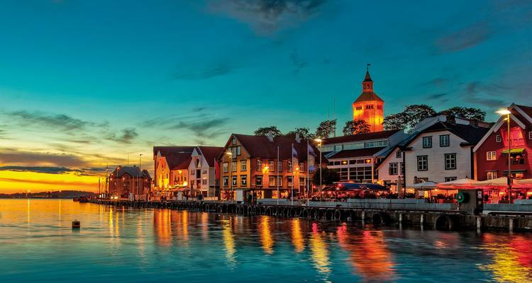 Las coloridas casas del frente de agua y la torre de la iglesia de Stavanger brillan bajo un atardecer vibrante con reflejos en aguas tranquilas.