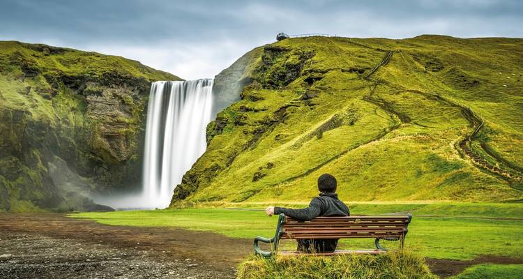 Eine Person sitzt auf einer Bank und genießt den Blick auf den Skogafoss-Wasserfall.