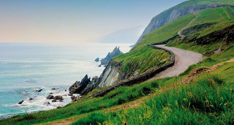 Die schmale Küstenstraße windet sich entlang steiler grüner Hänge über nebligen Atlantikgewässern auf dem Slea Head Drive.