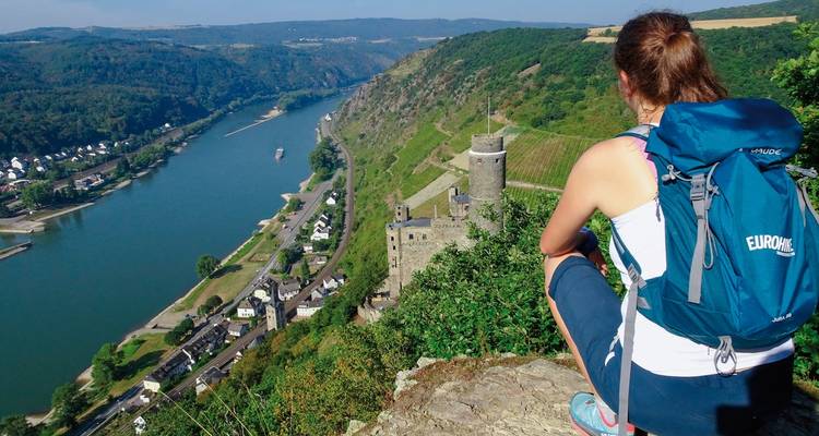 Un randonneur assis sur un rocher, profitant de la vue sur une rivière et un château.