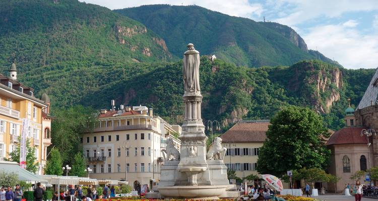 Statue sur une place de ville animée avec des montagnes en arrière-plan.