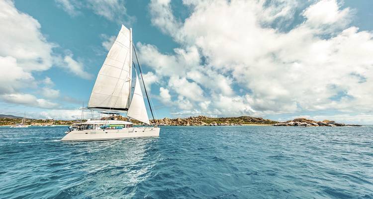 A sailboat on clear blue water with a rocky shoreline and partly cloudy sky.