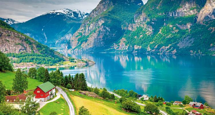 Spiegelglatter Fjord, umgeben von schneebedeckten Bergen, grünen Wäldern und einem roten Bauernhaus auf sanften Weiden in Norwegen.