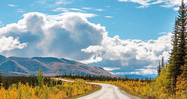 Vue panoramique d'une route sinueuse traversant un paysage montagneux avec un feuillage d'automne et un ciel nuageux.