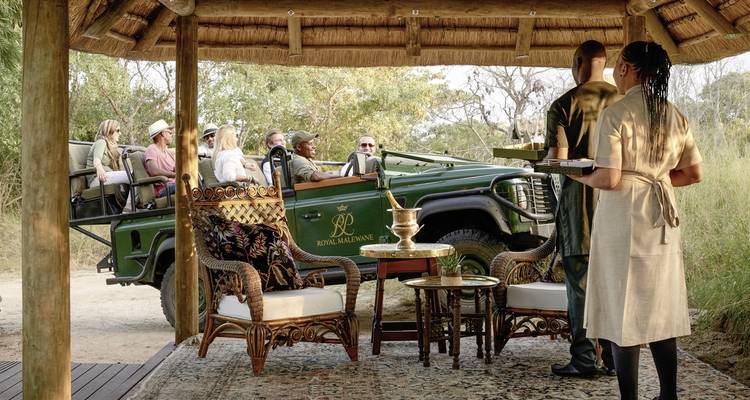 Touristen in einem Safari-Fahrzeug, das vor einer Lodge geparkt ist, mit Personal im Vordergrund, das serviert.