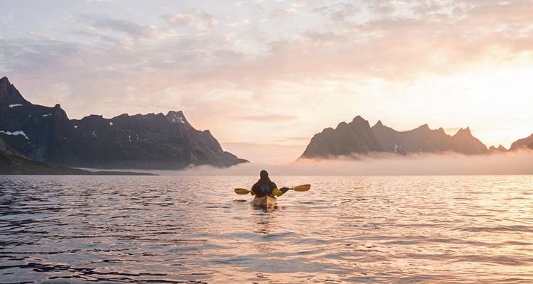 Kajakker die paddelt richting bergachtig landschap tijdens zonsondergang.