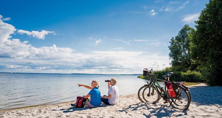 Zwei Menschen, die an einem sandigen Strand neben Fahrrädern sitzen und aufs Wasser blicken.
