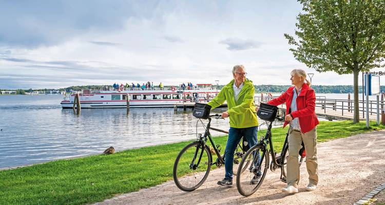 Zwei Personen mit Fahrrädern in der Nähe eines Sees mit einem Ausflugsboot.