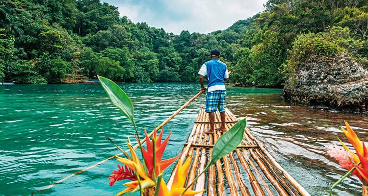 Person rafting on a bamboo raft on a scenic river surrounded by lush foliage.