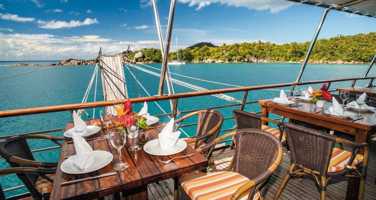 Outdoor dining area on a ship with a view of turquoise water and Seychelles landscape.