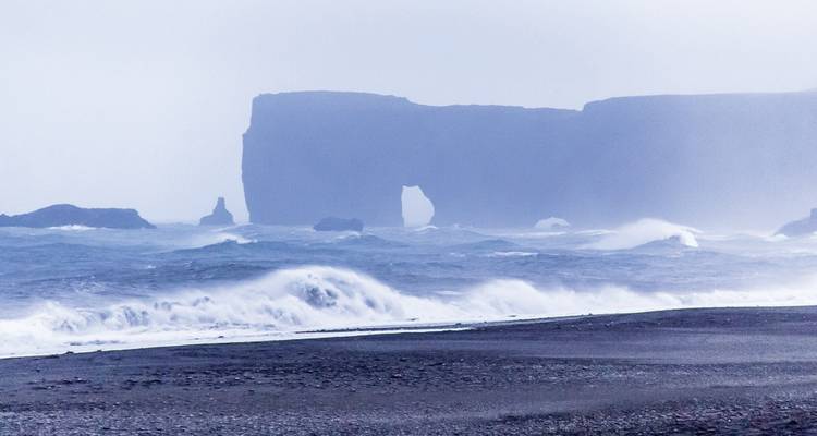 Une plage rocheuse avec des vagues qui se brisent et une arche naturelle en arrière-plan.