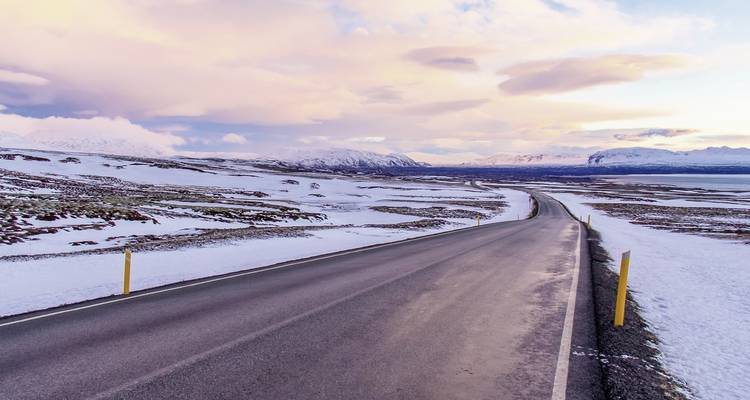 Une route enneigée menant vers des montagnes lointaines sous un ciel de coucher de soleil.