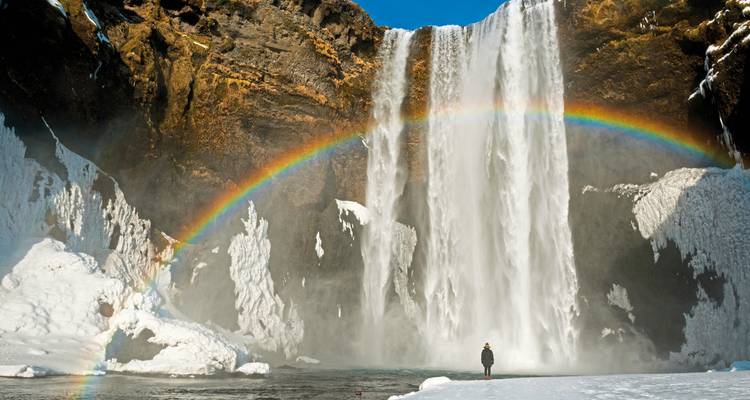Une personne debout à la cascade de Skógafoss avec un arc-en-ciel en vue.