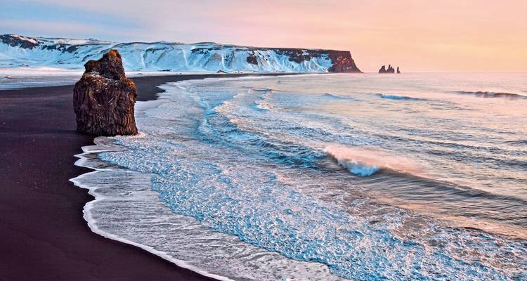 Une plage de sable noir avec des vagues et des falaises enneigées au coucher du soleil.