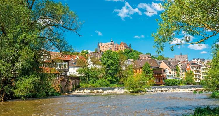 Village au bord de la rivière avec des bâtiments historiques et un château sur une colline.
