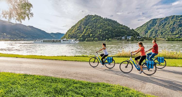 Trois cyclistes roulent le long d'une rivière avec un bateau de croisière et des collines.