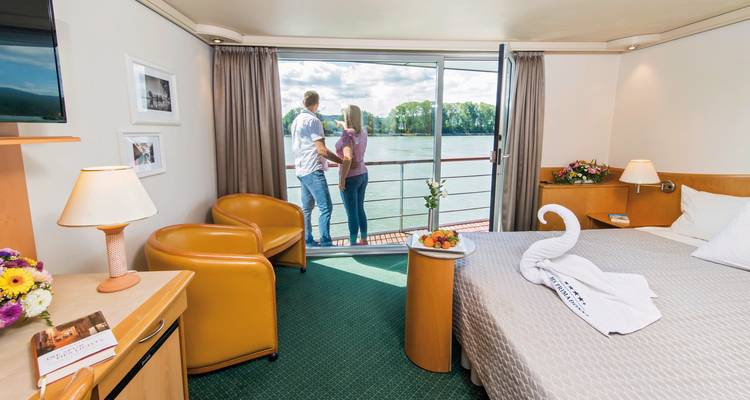Un couple debout près d'une fenêtre dans une cabine de bateau de croisière fluviale avec vue sur la rivière et le paysage lointain.