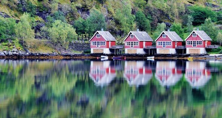Hellrote Häuser auf Stelzen, die sich im ruhigen Wasser eines Fjords spiegeln.