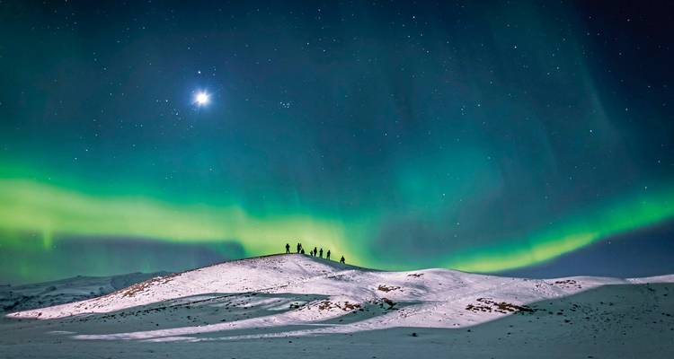 Aurore boréale au-dessus d'une colline enneigée avec des personnes en silhouette.